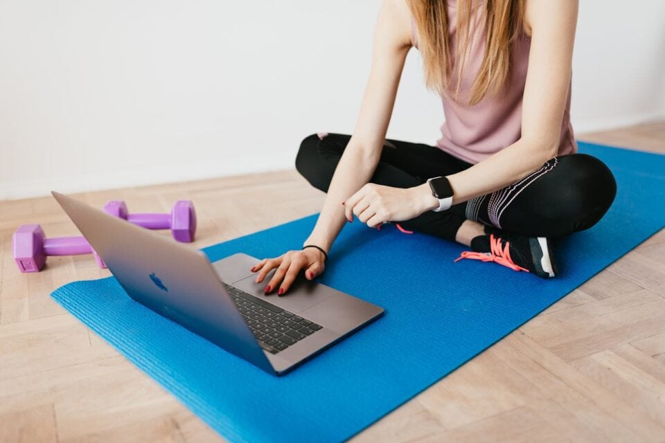 Woman on her laptop reading her program from her online fitness coach, sitting on a yoga mat next to some dumbells